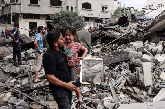 Graphic content / TOPSHOT - A man carries a crying child as he walks in front of a building destroyed in an Israeli air strike in Gaza City on October 7, 2023. Palestinian militants have begun a "war" against Israel which they infiltrated by air, sea and land from the blockaded Gaza Strip, Israeli officials said, a major escalation in the Israeli-Palestinian conflict